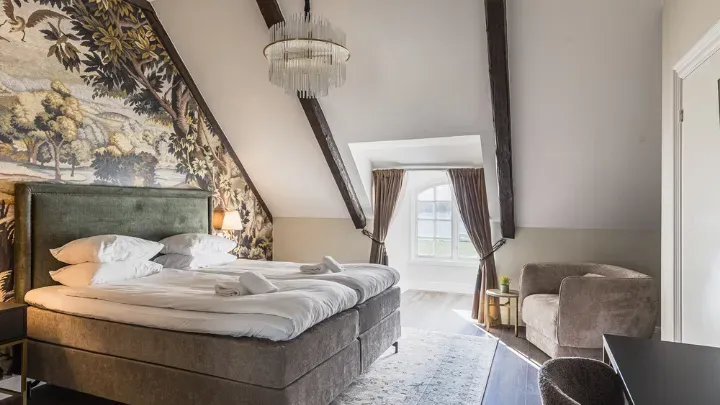 A made bed beneath exposed roof beams, with a crystal chandelier and seating beside the window.