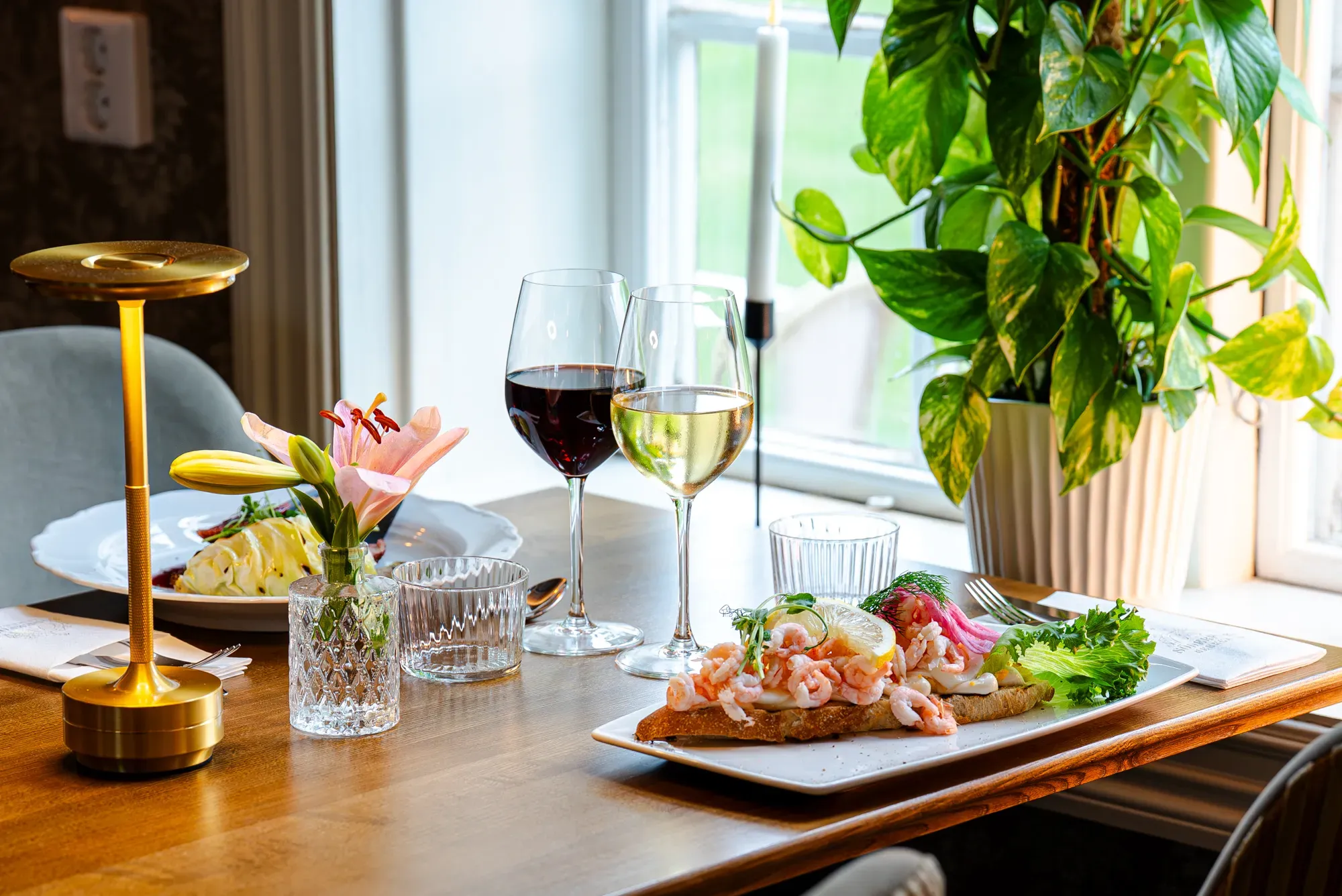 Open shrimp sandwiches, wine, and flowers set beside a restaurant window.