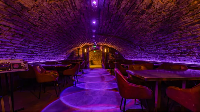 The cellar vault in purple light, with tables and chairs along the stone wall.